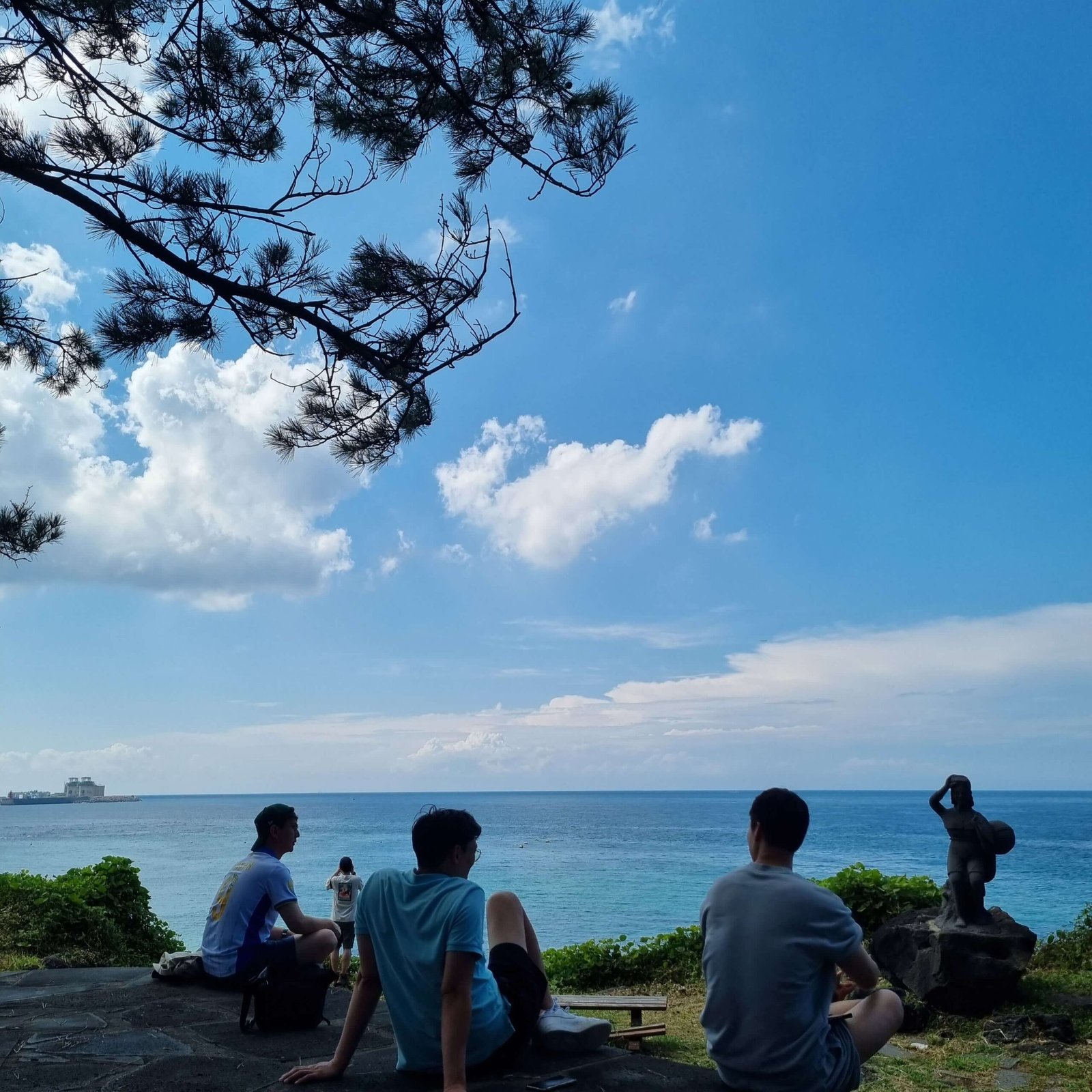 Friends relaxing by a Haenyeo statue with a view of the emerald ocean, capturing the essence of Jeju Island travel