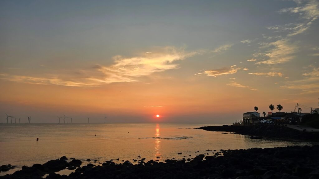 Coastal wind turbines and rocky shores under a beautiful orange sunset sky, a must-see during Jeju Island travel